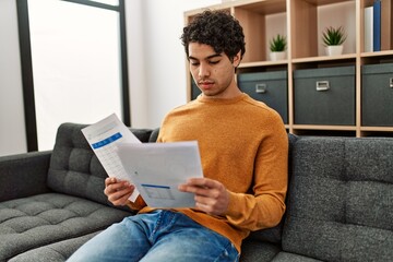 Canvas Print - Young hispanic man reading paperwork sitting on the sofa at home.