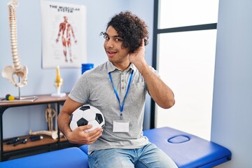 Wall Mural - Hispanic man with curly hair working as football physiotherapist smiling with hand over ear listening an hearing to rumor or gossip. deafness concept.