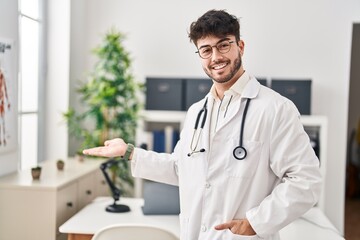 Wall Mural - Young hispanic man wearing doctor uniform doing welcome gesture with hand at clinic