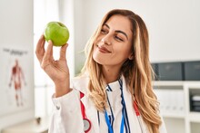 Young Blonde Woman Wearing Doctor Uniform Holding Apple At Clinic