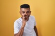 © Krakenimages.com - Young hispanic man standing over yellow background hand on mouth telling secret rumor, whispering malicious talk conversation