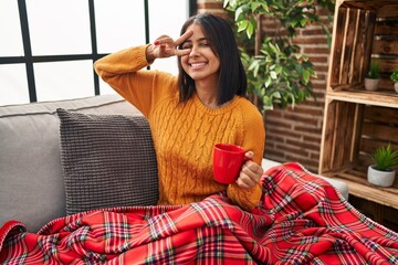 Wall Mural - Young hispanic woman sitting on the sofa drinking a coffee at home doing peace symbol with fingers over face, smiling cheerful showing victory