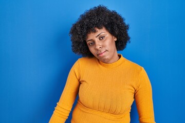 Poster - Black woman with curly hair standing over blue background looking sleepy and tired, exhausted for fatigue and hangover, lazy eyes in the morning.