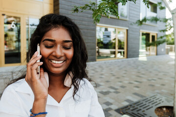 Wall Mural - Young brunette indian woman smiling and talking on cellphone outdoors