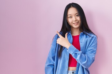 Sticker - Young asian woman standing over pink background cheerful with a smile on face pointing with hand and finger up to the side with happy and natural expression