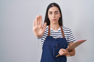 Wall Mural - Young brunette woman wearing professional waitress apron and clipboard with open hand doing stop sign with serious and confident expression, defense gesture