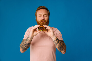 Wall Mural - Bearded man eating burger while standing isolated over blue background