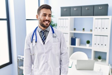 Wall Mural - Handsome hispanic man wearing doctor uniform and stethoscope at medical clinic looking away to side with smile on face, natural expression. laughing confident.