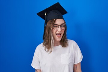 Wall Mural - Blonde caucasian woman wearing graduation cap winking looking at the camera with sexy expression, cheerful and happy face.