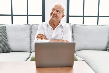 Sticker - Senior man using laptop at home sitting on the sofa smiling looking to the side and staring away thinking.