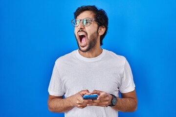 Canvas Print - Handsome latin man using smartphone typing message angry and mad screaming frustrated and furious, shouting with anger. rage and aggressive concept.