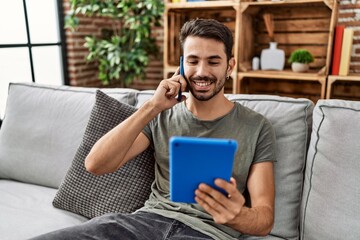 Wall Mural - Young hispanic man using touchpad and talking on the smartphone at home