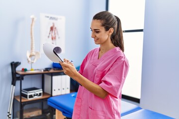 Canvas Print - Young hispanic woman wearing physiotherapist uniform writing on clipboard at physiotherpy clinic