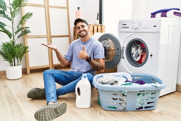 Poster - Young hispanic man putting dirty laundry into washing machine showing palm hand and doing ok gesture with thumbs up, smiling happy and cheerful