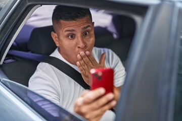 Canvas Print - Young hispanic man doing video call with smartphone in the car covering mouth with hand, shocked and afraid for mistake. surprised expression