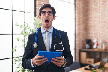 Canvas Print - Young hispanic man wearing doctor stethoscope at the office angry and mad screaming frustrated and furious, shouting with anger looking up.