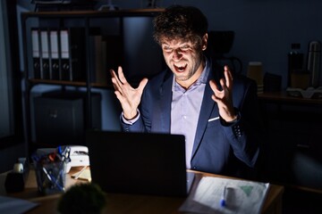 Canvas Print - Hispanic young man working at the office at night crazy and mad shouting and yelling with aggressive expression and arms raised. frustration concept.