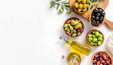 Different Olives In Bowls On White Concrete Background. Top View Of Olives, Olive Leaves And Bottle Of Olive Oil. Diet Food Concept. Banner.