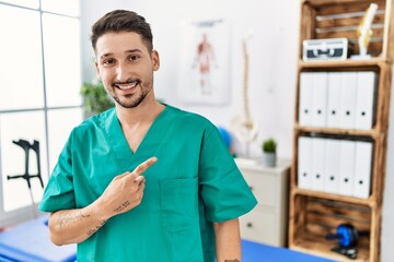 Canvas Print - Young physiotherapist man working at pain recovery clinic cheerful with a smile on face pointing with hand and finger up to the side with happy and natural expression