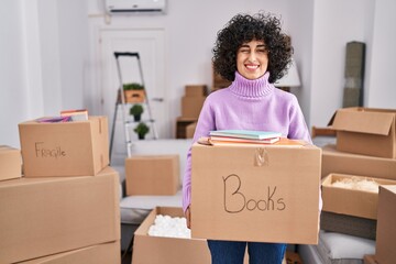 Canvas Print - Young brunette woman with curly hair moving to a new home holding cardboard box winking looking at the camera with sexy expression, cheerful and happy face.