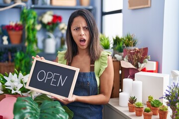 Canvas Print - Hispanic young woman working at florist with open sign in shock face, looking skeptical and sarcastic, surprised with open mouth