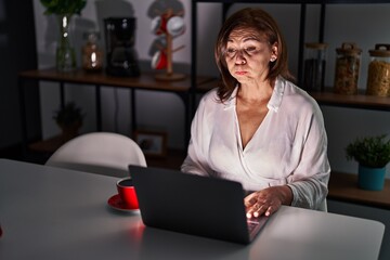 Sticker - Middle age hispanic woman using laptop at home at night puffing cheeks with funny face. mouth inflated with air, crazy expression.