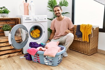 Poster - Young hispanic man putting dirty laundry into washing machine looking positive and happy standing and smiling with a confident smile showing teeth