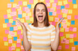 © sementsova321 - Overjoyed happy positive optimistic woman wearing striped t shirt standing against yellow wall covered with multicolored stickers, raised arms, screaming happily, being in good mood.