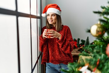 Poster - Young hispanic girl drinking coffee standing by christmas tree at home.