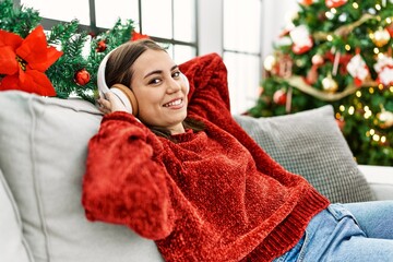 Sticker - Young hispanic girl listening to music sitting on the sofa by christmas tree at home.