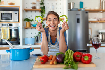 Photo of young woman smiling with green pepper slices while cooking salad with fresh vegetables in kitchen interior at home