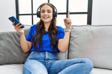 Wall Mural - Young brunette woman listening to music from smartphone using headphones smiling with an idea or question pointing finger with happy face, number one