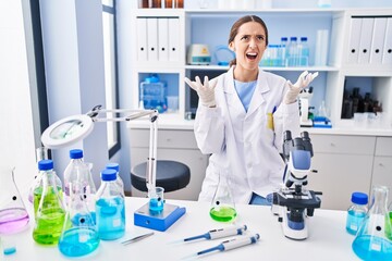 young brunette woman working at scientist laboratory crazy and mad shouting and yelling with aggress