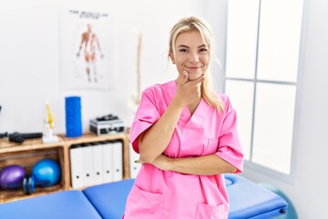 Wall Mural - Young caucasian woman working at pain recovery clinic looking confident at the camera with smile with crossed arms and hand raised on chin. thinking positive.