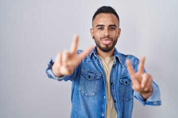 Poster - Young hispanic man standing over isolated background smiling with tongue out showing fingers of both hands doing victory sign. number two.