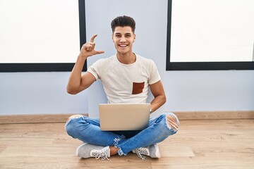 Wall Mural - Young hispanic man using laptop at home smiling and confident gesturing with hand doing small size sign with fingers looking and the camera. measure concept.