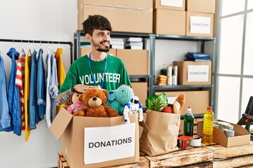 Wall Mural - Young hispanic man wearing volunteer uniform holding donations box with toys at charity center.