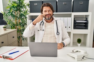 Canvas Print - Handsome middle age doctor man working at the clinic pointing with hand finger to face and nose, smiling cheerful. beauty concept