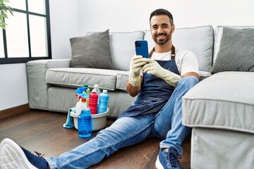 Canvas Print - Young hispanic man wearing apron and gloves using smartphone at home