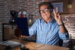 © Krakenimages.com - Middle age hispanic man using touchpad sitting on the table at night smiling looking to the camera showing fingers doing victory sign. number two.