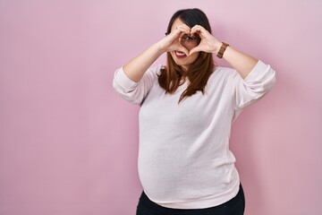 Poster - Pregnant woman standing over pink background doing heart shape with hand and fingers smiling looking through sign