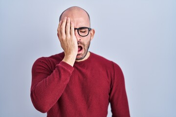 Wall Mural - Young bald man with beard standing over white background wearing glasses yawning tired covering half face, eye and mouth with hand. face hurts in pain.