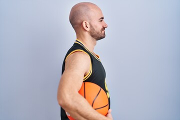 Wall Mural - Young bald man with beard wearing basketball uniform holding ball looking to side, relax profile pose with natural face and confident smile.