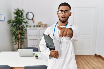 Sticker - Young indian man wearing doctor uniform and stethoscope pointing with finger surprised ahead, open mouth amazed expression, something on the front