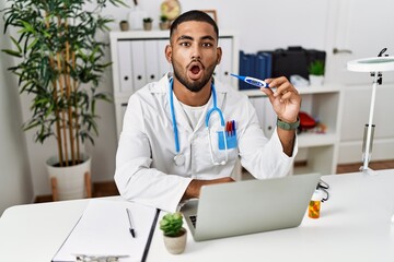 Poster - Young indian doctor holding thermometer scared and amazed with open mouth for surprise, disbelief face