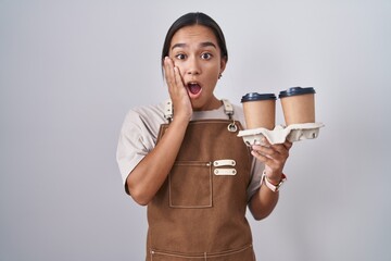 Wall Mural - Young hispanic woman wearing professional waitress apron holding coffee afraid and shocked, surprise and amazed expression with hands on face
