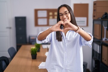 Wall Mural - Young hispanic woman at the office smiling in love doing heart symbol shape with hands. romantic concept.