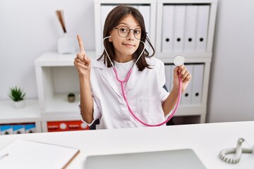 Poster - Young hispanic girl wearing doctor coat holding stethoscope smiling with an idea or question pointing finger with happy face, number one