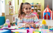 Adorable Hispanic Girl Student Sitting On Table Drawing On Paper At Kindergarten