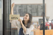 © David - Portrait of happy waitress standing at restaurant entrance. Portrait of young business Asian woman attend new customers in her coffee shop. Smiling small business owner showing open sign in her shop.